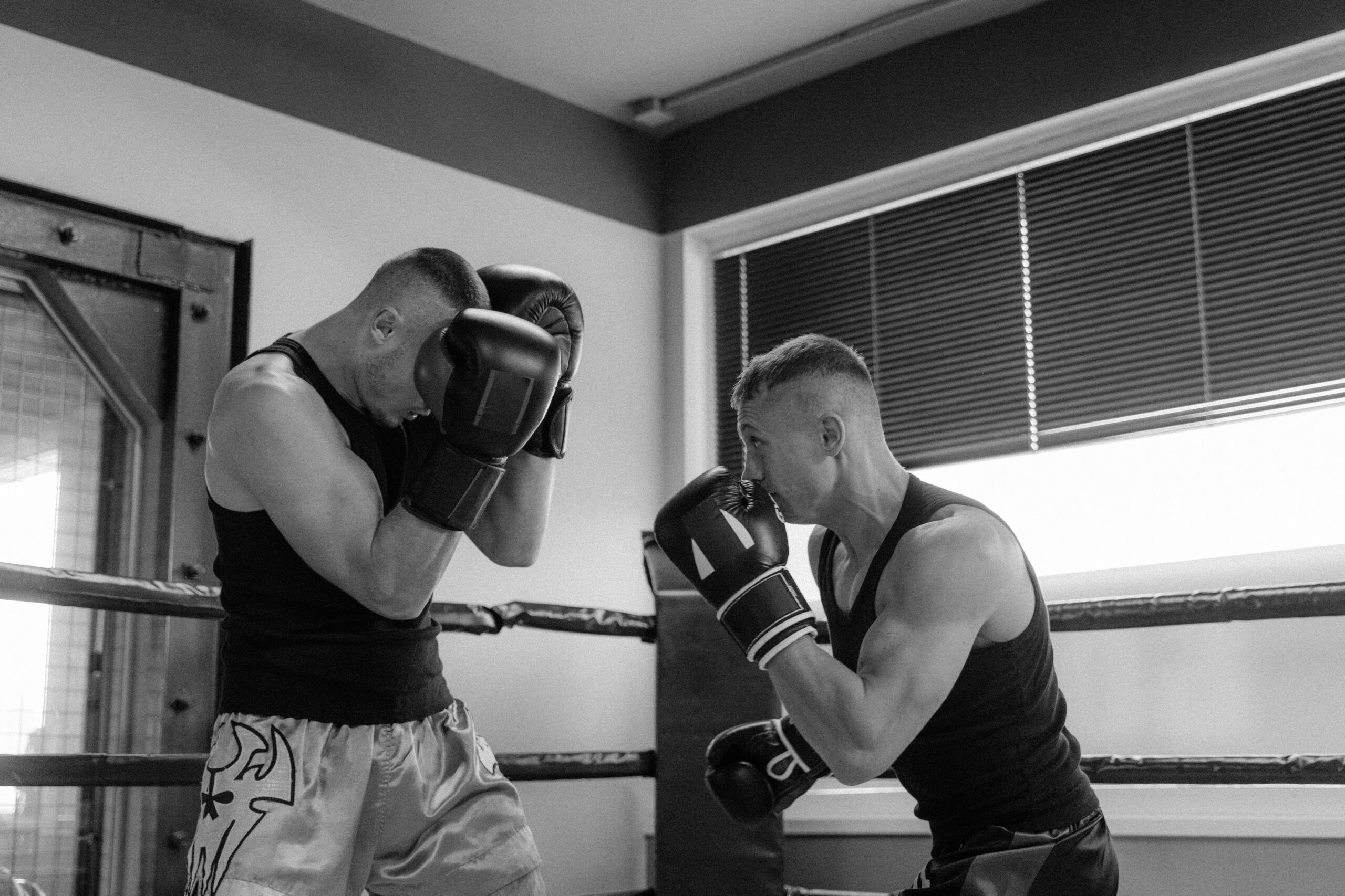 2 fighters sparring in a boxing gym. 1 fighter guarding his face with boxing gloves. The other bother is pucnhing.